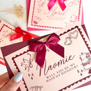 May include: Bridesmaid proposal cards in pink and burgundy, featuring decorative illustrations and text. The cards have a scalloped edge, a satin bow, and the names "April" and "Naomie". The text reads "Will you be my bridesmaid?" and "Will you be my maid of honour?"