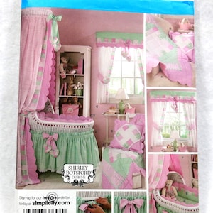 Op de afbeelding: Simplicity naaipatroon met een kinderkamer set met roze en groene gordijnen, een wieg en beddengoed. Het patroon bevat een wiegrok, gordijnen en een quilt. De tekst "SHIRLEY BOTSFORD DESIGNS" is zichtbaar.