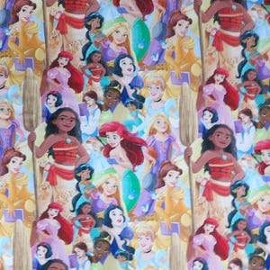 May include: A colorful fabric featuring a repeating pattern of various Disney princesses. The princesses are depicted in their iconic outfits, with a mix of skin tones and hair colors. The background is a light yellow.