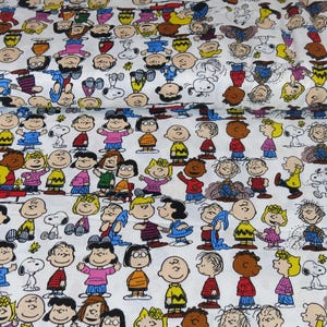 May include: White fabric featuring a repeating pattern of cartoon characters in various poses and outfits. The characters are depicted in a variety of colors, including yellow, red, blue, and brown. The fabric is rolled up.