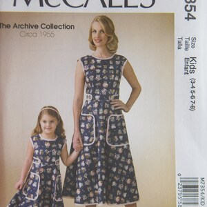 May include: McCall's sewing pattern M7354 featuring a mother and child in matching dresses. The dresses are navy blue with a whimsical print and pink trim. The pattern is from The Archive Collection, circa 1955, and includes sizes for kids.