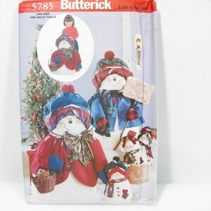 以下が含まれることがあります： 雪だるまの装飾が施されたButterickの裁縫パターン。雪だるまは、赤、青、チェック柄の帽子、スカーフ、コートを着ています。1つの雪だるまは「Happy Holidays from Our Family!」と書かれた看板を持っています。