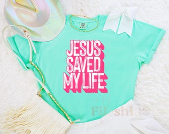 May include: A turquoise blue t-shirt with the text "Jesus Saved My Life" in pink lettering. The shirt is made of a soft, comfortable material and has a relaxed fit.