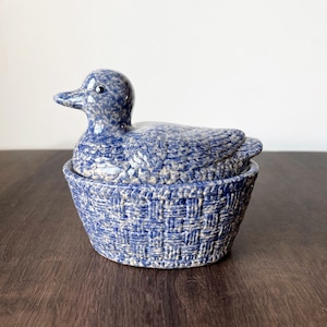 May include: A blue and white ceramic duck figurine atop a basket-shaped container. The duck has a speckled pattern and is perched on a basket with a woven design. The piece is likely a decorative item.