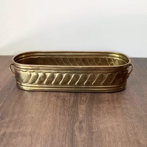 May include: A rectangular brass planter with a decorative leaf pattern around the sides and small handles. The planter is empty and sits on a wooden surface. The brass has a warm, golden hue.