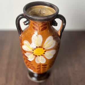 May include: A hand-painted ceramic vase with a brown base and body, featuring a large white and yellow flower design. The vase has two black handles and a dark brown rim. The vase is approximately 8 inches tall.