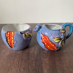 May include: A blue ceramic creamer and sugar bowl set. The set features a design of red and yellow accents. The creamer has a handle and spout. The sugar bowl is round. The set is on a wooden surface.