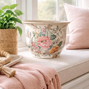 May include: Cream-colored ceramic planter with a floral design featuring pink and orange roses. The planter is round with a wide rim and sits on a window seat. A pink throw blanket with tassels is draped nearby.