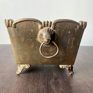 May include: Antique brass planter with a rectangular shape and ornate details. Features a lion's head with a ring handle and decorative feet. The planter has a weathered, aged appearance, suggesting it is vintage.