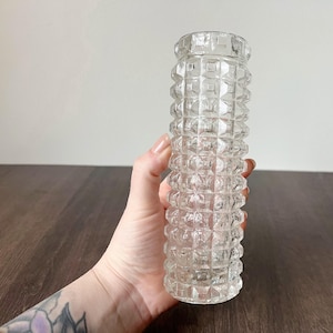 May include: A clear glass vase with a textured, geometric pattern. The vase is cylindrical and has a series of raised squares along its length. The vase is held in a hand, and the background is a dark wooden surface.