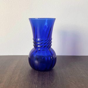 May include: A cobalt blue glass vase with a bulbous base and a textured, twisted design around the neck. The vase has a flared top and sits on a dark wooden surface. The vase is empty.