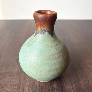 May include: A small, handcrafted ceramic vase with a rounded body and a narrow neck. The vase features a two-tone color scheme, with a light green body and a dark brown rim. The vase is approximately 5 inches tall.