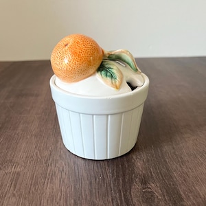 May include: White ceramic sugar bowl with a lid featuring an orange and green leaves. The bowl has vertical ribbed detailing. The orange is textured and has a realistic appearance. A small spoon-sized opening is visible.