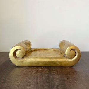 May include: A gold-colored, scroll-shaped decorative tray. The tray has a rectangular base with a circular indentation in the center. The ends of the tray curl upwards in a scroll design. The tray is made of a solid material.