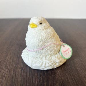 May include: A white, textured chick figurine with a yellow beak and black eyes, sitting on a white nest. A pink string and a tag that reads "Easter 1994" are attached. The figurine is likely a decorative Easter item.