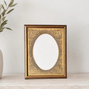 May include: A rectangular gold picture frame with an oval opening. The frame has ornate detailing around the oval and on the outer edges. A white vase with a sprig of greenery is in the background.