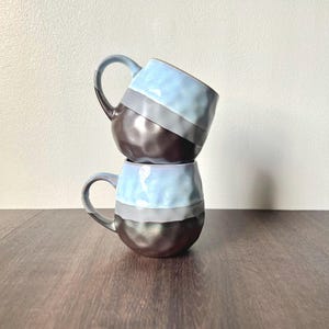 May include: Two stacked ceramic mugs. The mugs have a unique design with a light blue top, a gray middle section, and a dark brown bottom. Each mug has a curved handle for easy gripping. The mugs are on a wooden surface.