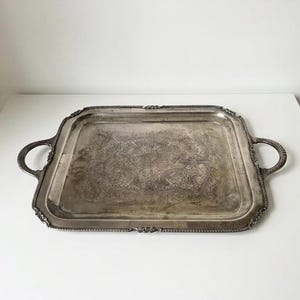 May include: A rectangular silver-toned serving tray featuring ornate, embossed floral patterns across the center surface. The tray includes two circular side handles and a decorative beaded border along the edges, showing a tarnished metallic finish.