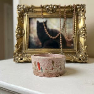 May include: A small, cylindrical, light pink concrete bowl with a speckled texture featuring scattered red and green paint accents along the exterior base. The bowl has a smooth, flat rim and a solid, sturdy construction.