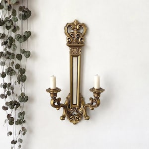 May include: A gold-colored wall sconce featuring an elongated vertical frame with a central mirror panel, topped with a decorative crown motif. Two curved arms extend from the base, each holding a white pillar candle in a circular holder.