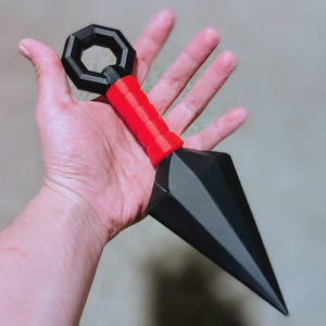 May include: A black and red kunai knife with a geometric design. The handle is red and cylindrical, leading to a black, faceted blade. The handle has a black, octagonal ring at the top.