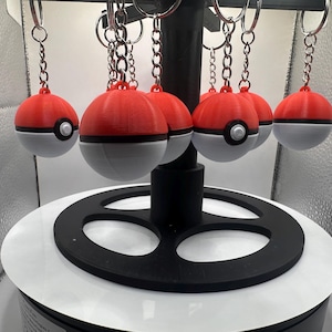 May include: Red and white ball-shaped keychains with a black stripe and a white button. The keychains are hanging from silver chains and are displayed on a black and white stand. The keychains are a popular item.