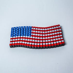 May include: A stylized American flag made of small, rectangular blocks. The flag features blue blocks with white stars in the upper left corner, and red and white stripes. The blocks are arranged to give a wavy appearance.