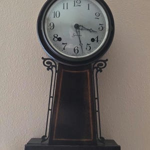 May include: A wall-mounted Sessions clock with a dark wood frame, a round white face featuring black Arabic numerals, and a brass eagle finial at the top. The lower section contains a rectangular glass panel displaying a landscape painting of a cabin.