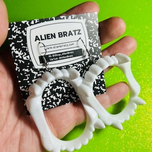 Pode incluir: Brincos de plástico branco em forma de dentes. Os brincos estão pendurados em ganchos prateados. Os brincos estão sobre um fundo preto e branco com o texto "Alien Bratz" e um endereço de site.