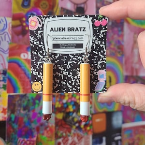 Pode incluir: Um par de brincos pendentes em forma de cigarros. Os brincos são amarelos com uma faixa vermelha e uma ponta branca. Os brincos estão pendurados em ganchos prateados.
