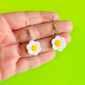 Op de afbeelding: Een paar witte bloemvormige oorbellen met een gele kern. De oorbellen hebben zilverkleurige haken. De oorbellen worden in een hand gehouden tegen een felgroene achtergrond.