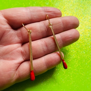 Pode incluir: Um par de brincos dourados em forma de fósforos. Cada brinco tem um corpo longo e fino de cor dourada com uma ponta vermelha. Os brincos são exibidos em um fundo verde brilhante.