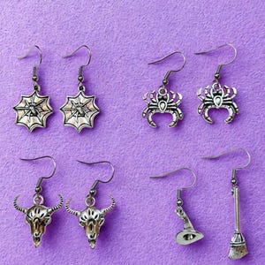 Pode incluir: Uma coleção de brincos prateados com tema de Halloween. Os designs incluem teias de aranha, caranguejos, crânios de touro, um chapéu de bruxa e uma vassoura. Os brincos são exibidos em um fundo roxo, mostrando seus detalhes.