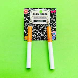 Pode incluir: Um par de brincos de argola prateados em forma de cigarros com um design branco e laranja. Os brincos são exibidos em um caderno preto e branco com o texto "Alien Bratz" e um endereço de site.