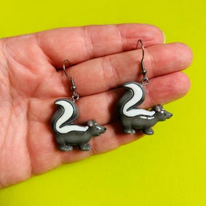 Pode incluir: Um par de brincos de doninha. Cada brinco apresenta um doninha cinzento com uma risca e cauda brancas. As doninhas estão presas a ganchos de orelha prateados. O fundo é amarelo brilhante.