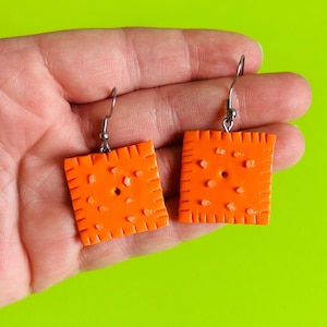 Pode incluir: Um par de brincos quadrados laranja que se assemelham a bolachas. Cada brinco tem uma superfície texturizada com pequenos detalhes em relevo e um único orifício. Pendurados em ganchos de orelha prateados.