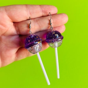 Pode incluir: Um par de brincos pendentes em forma de pirulitos. A parte superior é uma esfera roxa translúcida preenchida com glitter e confetes em forma de estrela, enquanto a parte inferior é uma esfera transparente com flocos prateados num palito branco.