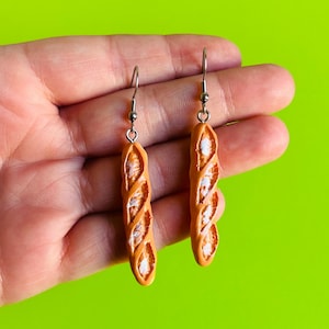 May include: A pair of dangle earrings featuring miniature baguette bread charms. Each charm is golden-brown with textured, scored crust details and white accents, suspended from silver-toned metal fishhook wires for pierced ears.