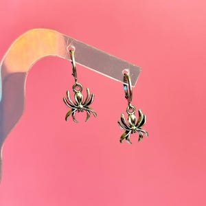 Op de afbeelding: Een paar zilverkleurige oorringen met bungelende spinnenhangers. Elke oorbel bestaat uit een kleine ronde ring met een gedetailleerde, achtpotige spinnenhanger die aan de onderkant is bevestigd, met een metalen afwerking.
