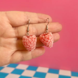 Pode incluir: Um par de brincos pendentes com pingentes realistas e texturizados em forma de framboesa rosa claro. Cada pingente está suspenso por um gancho de metal prateado simples. Os pingentes de framboesa têm uma aparência macia e aveludada.