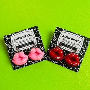 Pode incluir: Dois pares de brincos em forma de lábios, um em rosa claro e outro em vermelho, exibidos em cartões com padrão em preto e branco. Os cartões apresentam o texto "ALIEN BRATZ" e informações do site e das redes sociais. O fundo é verde brilhante.