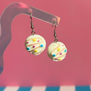 Op de afbeelding: Een paar hangende oorbellen met ronde, witte, getextureerde bedels die lijken op bolletjes ijs. Elke bedel is versierd met dunne roze glazuurwervelingen, kleine gele en blauwe stervormige spikkels en kleine roze ronde stippen.