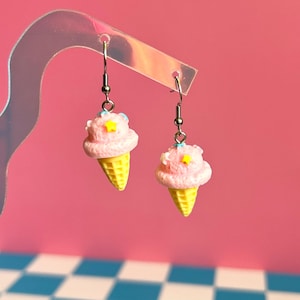 Op de afbeelding: Een paar bungelende oorbellen met miniatuur ijshoorntjes met roze bollen, gele wafelhoorntjes en kleine gele stervormige spikkels, hangend aan zilverkleurige metalen haakjes bevestigd aan een doorzichtige, gebogen displaystandaard.