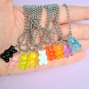Pode incluir: Uma coleção de colares de ursinhos de goma coloridos. Cada colar apresenta um pingente de ursinho de goma de cor diferente, incluindo preto, rosa, amarelo, laranja, branco, roxo e azul. Os pingentes são presos a uma corrente de bolinhas prateada.