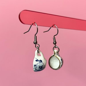 Pode incluir: Brincos pendurados com mini chávenas de chá de porcelana. Um brinco é uma chávena, o outro é uma chávena de chá. Ambos são brancos com desenhos florais azuis e acabamentos dourados, suspensos por ganchos prateados. O fundo é rosa liso.