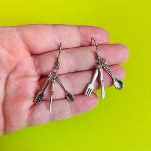 Pode incluir: Brincos pendurados prateados com pingentes em miniatura de garfo, colher e faca. Cada brinco tem um pequeno gancho para orelhas furadas. Os brincos são exibidos em um fundo amarelo-esverdeado brilhante.