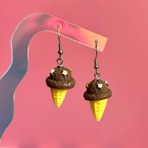 Op de afbeelding: Een paar hangende oorbellen met miniatuur chocolade-ijsbolletjes in gele wafelhoorntjes. Het bruine ijs is versierd met kleine, veelkleurige stervormige spikkels. De bedels hangen aan zilverkleurige metalen oorhaken.