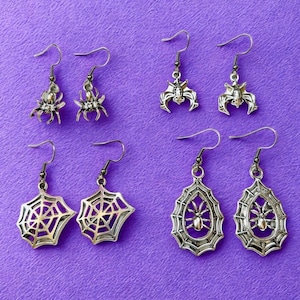 Pode incluir: Uma coleção de brincos prateados com tema de Halloween. Os brincos apresentam desenhos de aranha, morcego, teia de aranha e aranha na teia. Os brincos são exibidos em um fundo roxo.