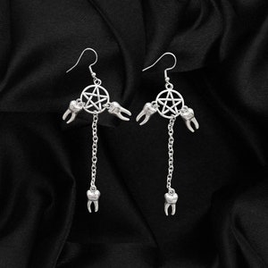 May include: Silver earrings with a pentagram charm and dangling teeth. The earrings are on a black fabric background.