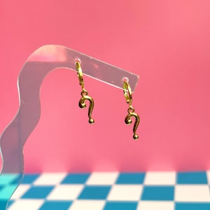 Op de afbeelding: Een paar goudkleurige oorringen, elk voorzien van een bungelende vraagtekenhanger. De oorbellen hangen aan een doorzichtige, golvende displaystandaard, waardoor de metallic afwerking en de kenmerkende gebogen vorm van de hangers zichtbaar zijn.
